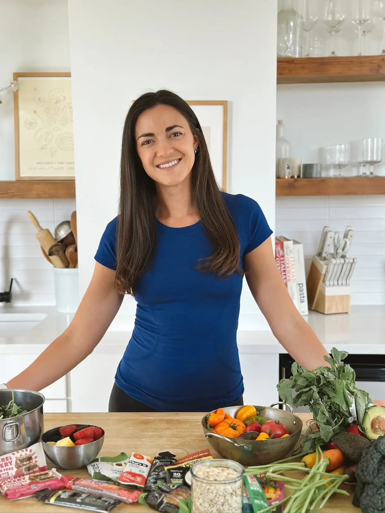 A dietitian in a blue shirt smiles in a kitchen, standing behind a table of fresh fruits, veggies, oats, and snacks used to teach swim families about nutrition on Eat Swim Win. Eat Swim Win